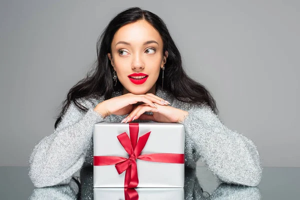 Glückliche Frau mit roten Lippen, die in der Nähe der Geschenkschachtel wegschaut, isoliert auf grau — Stockfoto