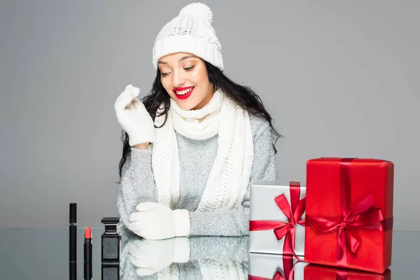 Happy woman in winter outfit looking at decorative cosmetics near presents isolated on grey — Stock Photo