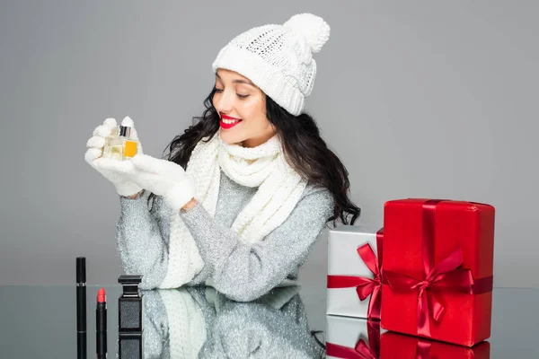 Glückliche Frau im winterlichen Outfit mit Parfüm in der Nähe von Geschenken isoliert auf grau — Stockfoto