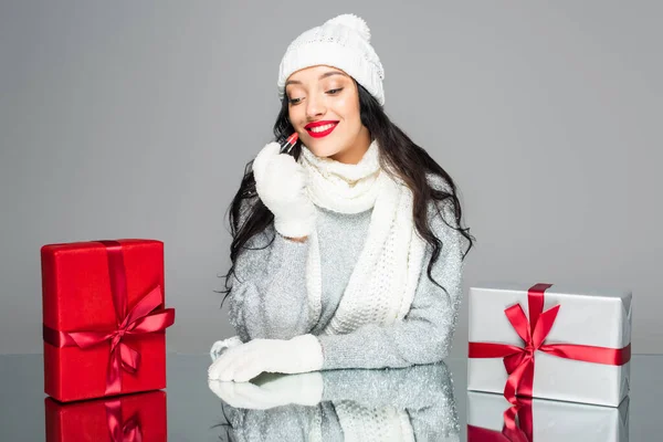 Щаслива жінка в зимовому вбранні тримає помаду біля подарунків ізольовано на сірому — стокове фото