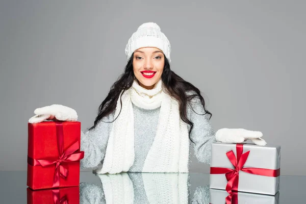 Щаслива жінка в зимовому вбранні дивиться на камеру біля подарунків ізольовано на сірому — стокове фото