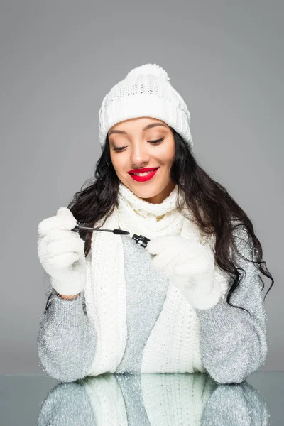 Fröhliche und brünette Frau im Winter-Outfit mit Wimperntusche isoliert auf grau — Stockfoto