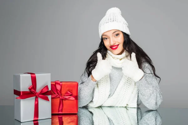 Fröhliche und brünette Frau im Winter-Outfit zeigt Daumen hoch in der Nähe von Geschenken isoliert auf grau — Stockfoto
