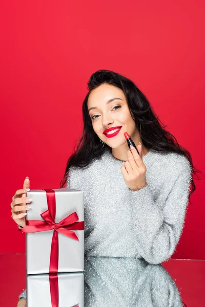 Femme heureuse en pull tenant rouge à lèvres près enveloppé présent isolé sur rouge — Photo de stock