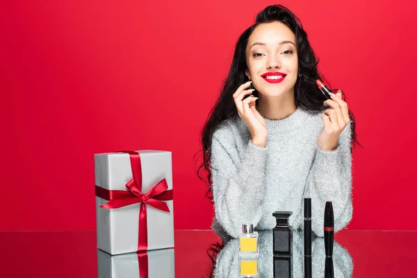 Cheerful woman in sweater holding lipstick near decorative cosmetics, bottle with perfume and wrapped present isolated on red — Stock Photo