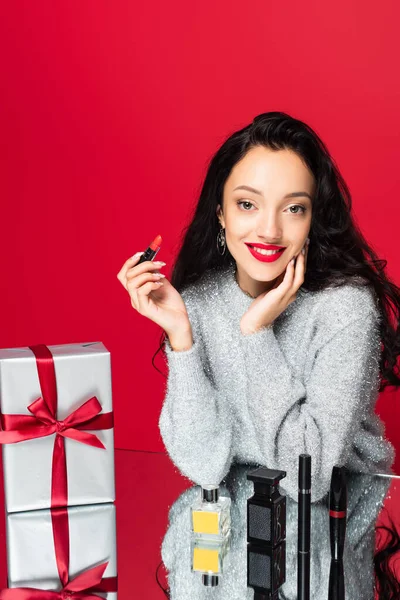 Smiling woman in sweater holding lipstick near decorative cosmetics and wrapped present isolated on red — Stock Photo