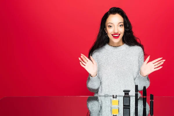 Smiling woman in sweater gesturing near decorative cosmetics and bottles with perfume isolated on red — Stock Photo