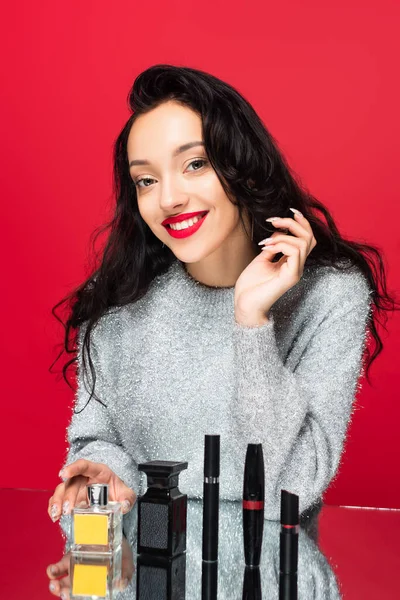 Piacere donna in maglione guardando la fotocamera vicino cosmetici decorativi e bottiglie con profumo isolato su rosso — Foto stock