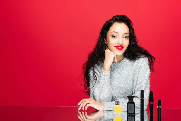 Jeune femme heureuse en pull regardant la caméra près des cosmétiques décoratifs et des bouteilles avec du parfum isolé sur rouge — Photo de stock