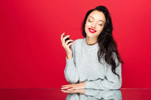 Young pleased woman with closed eyes holding bottle and smelling perfume isolated on red — Stock Photo