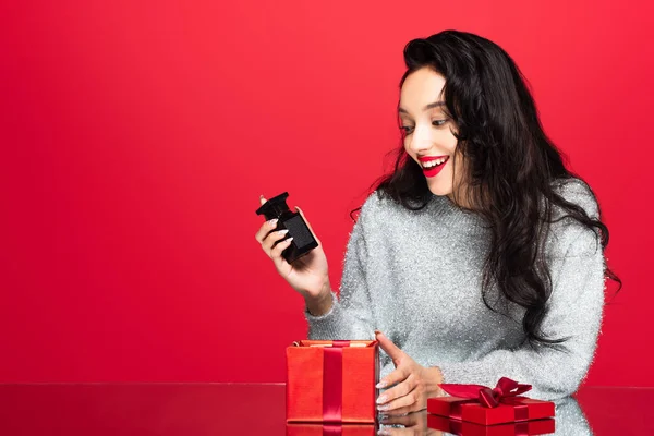 Glückliche Frau hält Flasche mit Parfüm in der Nähe Geschenkbox isoliert auf rot — Stockfoto