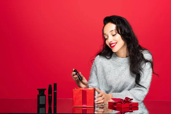 Glückliche Frau nimmt Lippenstift aus Geschenkbox in der Nähe von dekorativer Kosmetik auf Rot — Stockfoto