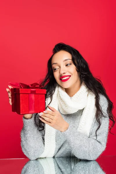 Fröhliche und brünette Frau im Pullover sieht Weihnachtsgeschenk isoliert auf rot — Stockfoto