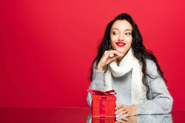 Fröhliche und brünette Frau im Pullover, die in der Nähe des Weihnachtsgeschenks wegschaut, isoliert auf rot — Stockfoto