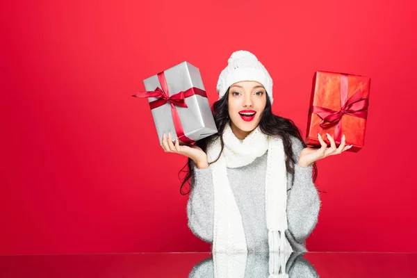 Mujer alegre en sombrero y bufanda caliente sosteniendo regalos aislados en rojo - foto de stock