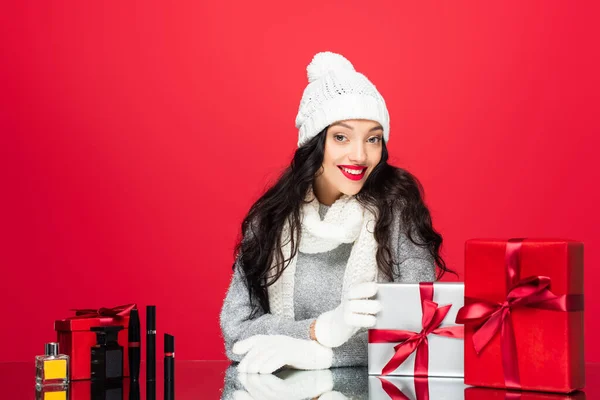 Femme heureuse en chapeau, gants et écharpe chaude près de cadeaux de Noël et cosmétiques décoratifs isolés sur rouge — Photo de stock