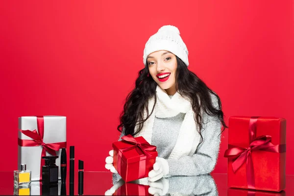 Улыбающаяся женщина в шляпе, перчатках и теплом шарфе возле рождественских подарков и декоративной косметики, изолированной на красном — стоковое фото