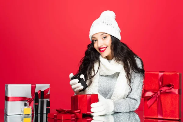 Joyful woman in hat, gloves and warm scarf holding bottle with perfume near christmas presents and decorative cosmetics isolated on red — Stock Photo
