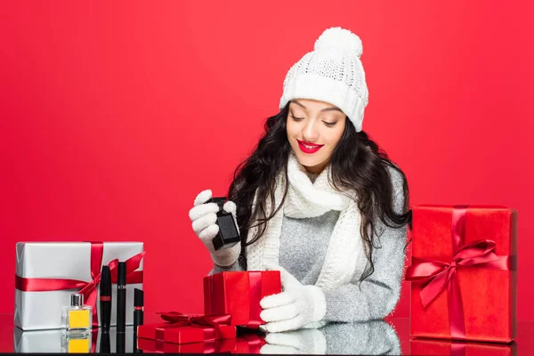 Femme joyeuse en tenue d'hiver tenant bouteille avec parfum près de cadeaux de Noël et cosmétiques décoratifs isolés sur rouge — Photo de stock