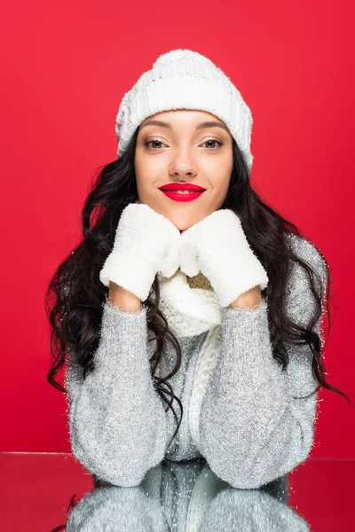 Pleased and brunette woman in hat and gloves looking at camera isolated on red — Stock Photo