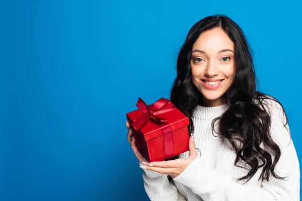 Allegra donna bruna in maglione a maglia che tiene il Natale presente isolato sul blu — Foto stock