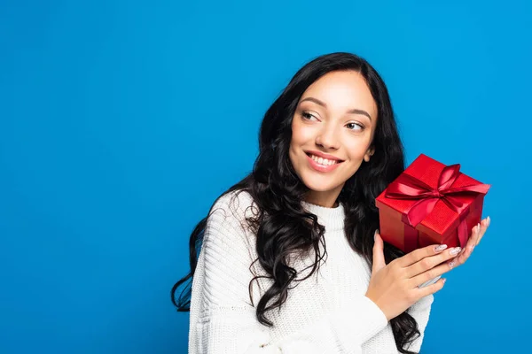 Glückliche brünette Frau im Strickpullover hält Weihnachtsgeschenk in der Hand und schaut isoliert auf blauem Grund weg — Stockfoto