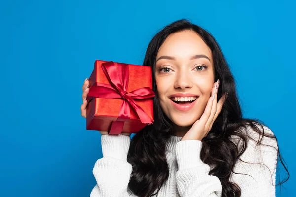 Happy brunette woman in knitted sweater holding christmas present isolated on blue — Stock Photo