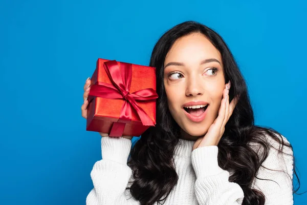 Aufgeregte brünette Frau im Strickpullover mit Weihnachtsgeschenk auf blauem Grund — Stockfoto