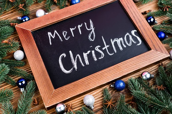 Kreidetafel mit frohem weihnachtlichen Schriftzug, neben Tannenzweigen, Kugeln und Anissternen auf braunem Holzgrund — Stockfoto