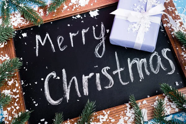 Vista de cerca de pizarra con tipografía feliz Navidad cerca de ramas de pino, pequeña caja de regalo y nieve artificial - foto de stock