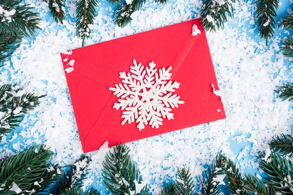 Top view of decorative snowflake on red envelope near pine branches and artificial snow on blue background — Stock Photo