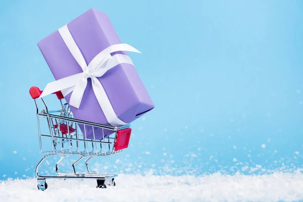 Pequeña caja de regalo en carrito de compras y nieve artificial sobre fondo azul, concepto de año nuevo - foto de stock