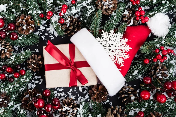 Vue de dessus des branches d'épinette avec neige artificielle, boîte cadeau près du chapeau de Père Noël et flocon de neige décoratif sur fond noir — Photo de stock