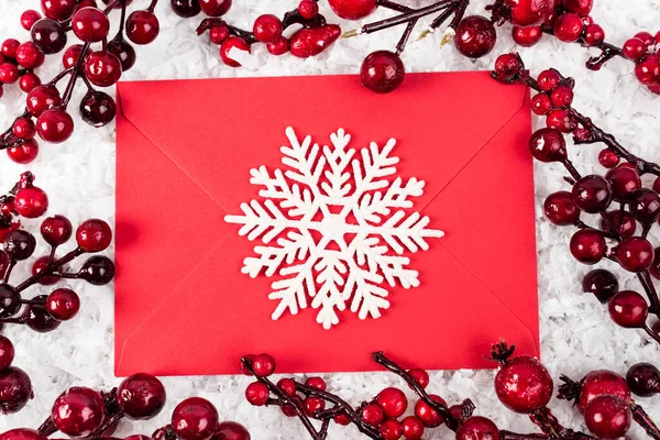 Вид зверху на декоративну сніжинку на червоному конверті біля гілок зі штучними ягодами на білому фактурному фоні, концепція нового року — стокове фото
