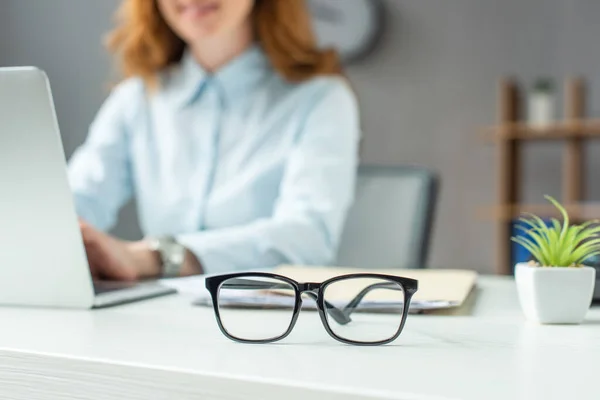 Cropped view of businesswoman typing on laptop, while sitting at table with eyeglasses on blurred background — Stock Photo