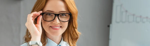 Mujer de negocios sonriente tocando el marco de las gafas y mirando a la cámara con el rotafolio borroso en el fondo, pancarta - foto de stock