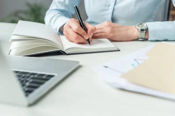 Vista recortada de la mujer de negocios escribiendo en el cuaderno, mientras está sentado en el lugar de trabajo en primer plano borrosa - foto de stock