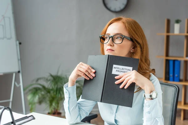 Nachdenklicher Anwalt hält Buch mit Schriftzug zum geistigen Eigentum, während er am Arbeitsplatz auf verschwommenem Hintergrund wegschaut — Stockfoto