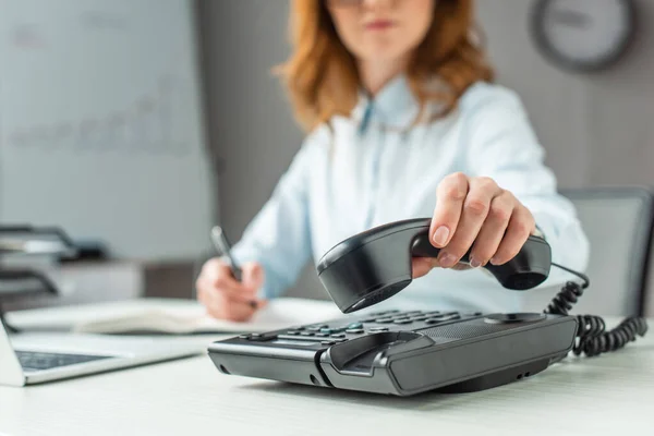 Cropped view of businesswoman putting handset on landline telephone, while sitting at workplace on blurred background — Stock Photo