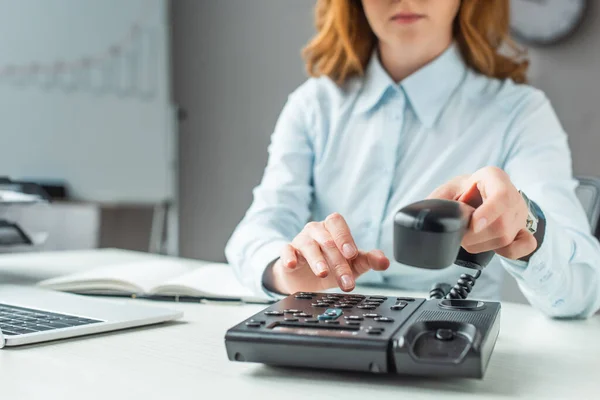 Ausgeschnittene Ansicht einer Geschäftsfrau mit Handywählnummer am Festnetztelefon am Arbeitsplatz auf verschwommenem Hintergrund — Stockfoto