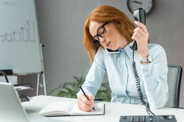 Mujer de negocios pelirroja con auricular hablando por teléfono móvil, mientras escribe en un cuaderno en el lugar de trabajo en un fondo borroso - foto de stock