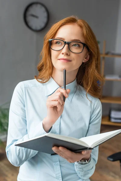 Donna d'affari positiva con il pensiero del taccuino, mentre distoglie lo sguardo con ufficio offuscato sullo sfondo — Foto stock