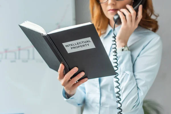 Vista recortada de la mujer de negocios sosteniendo libro con letras de propiedad intelectual, mientras habla por teléfono fijo sobre fondo borroso - foto de stock