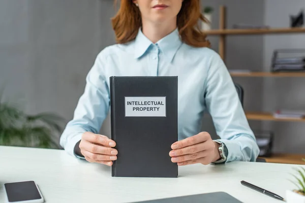 Ausgeschnittene Ansicht einer Rechtsanwältin, die ein Buch mit Schriftzug zum geistigen Eigentum zeigt, während sie am Arbeitsplatz auf verschwommenem Hintergrund sitzt — Stockfoto