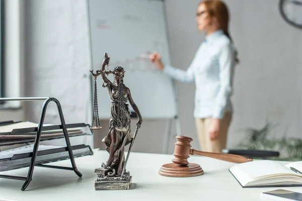 Estatuilla de Themis con mazo y bloque de madera en la mesa con abogado femenino borroso de pie cerca de flipchart en el fondo - foto de stock