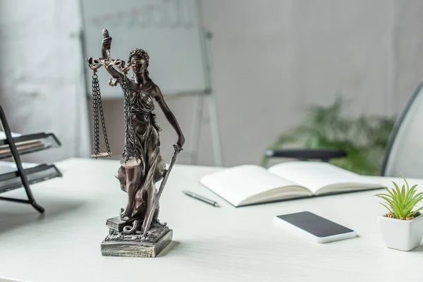 Themis Figur mit Handy und leerem Notizbuch auf Tisch mit unscharfem Flipchart im Hintergrund — Stockfoto