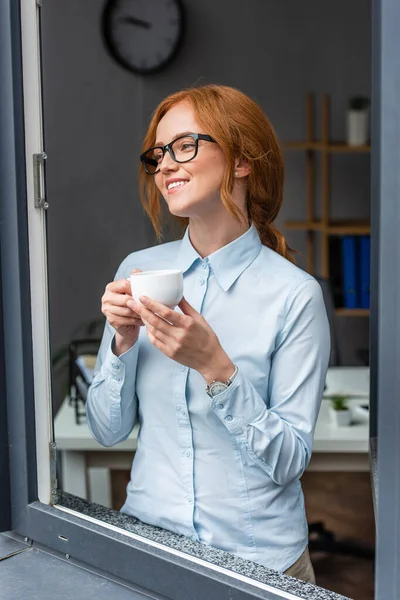 Glückliche rothaarige Geschäftsfrau mit Kaffeetasse, die wegschaut, während sie am Fenster steht, mit verschwommenem Büro im Hintergrund — Stockfoto