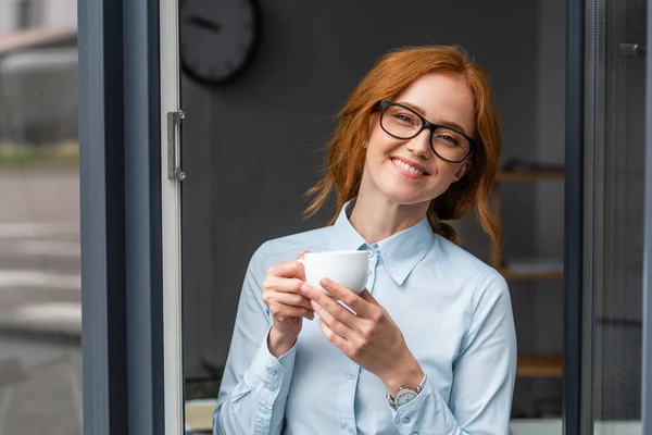 Feliz pelirroja empresaria con taza de café mirando a la cámara en la ventana - foto de stock