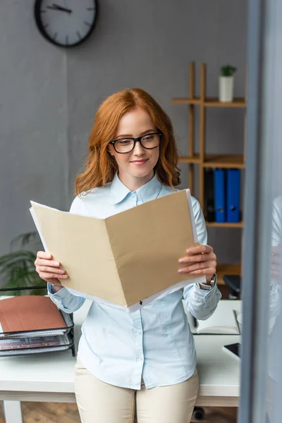 Lächelnde Geschäftsfrau blickt auf Ordner mit Papierblättern, während sie sich am Arbeitsplatz mit verschwommenem Fenster im Vordergrund lehnt — Stockfoto