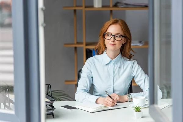 Positive Geschäftsfrau schreibt in Notizbuch, während sie am Arbeitsplatz mit verschwommenem Fenster im Vordergrund wegschaut — Stockfoto